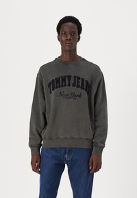 Donkergrijze sweatshirt met ronde hals, voorzien van een gestructureerd "TOMMY JEANS" logo in zwart, gecombineerd met blauwe spijkerbroeken. Casual fit met geribde manchetten.