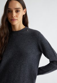 Maglione grigio carbone realizzato in morbido tessuto a maglia. Presenta un collo a coste, maniche lunghe e un piccolo dettaglio metallico sulla spalla.