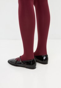 Zwarte gepatineerde leren schoenen met een t-bandontwerp, gecombineerd met bordeauxrode opake panty's. De zolen zijn plat en de neuzen zijn lichtjes puntschoen.