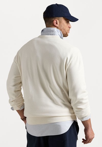 Kremna majica s crew vratom, z ribanim manžetami in robom, je plastena preko svetlo modre in bele črtaste srajce. Temno modra kapa dopolnjuje kombinacijo.