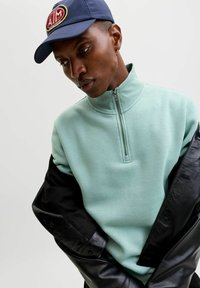 Felpa verde menta con collo alto e dettaglio con zip, indossata sotto una giacca di pelle nera, abbinata a un berretto blu navy con logo.