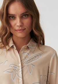 Beige Hemd mit Knöpfen und floralem sowie blättermuster in hellgrau. Verfügt über einen spitzen Kragen und eine glatte, glänzende Stoffoberfläche.
