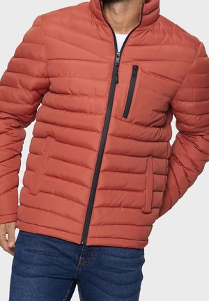 Man in een roestkleurige gewatteerde jas met zwarte ritsdetails en donkerblauwe jeans, poseert met de linkerhand in zijn zak.