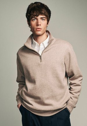 Sudadera beige con cremallera y alto cuello, de textura suave y mangas largas. Debajo, se asoma ligeramente una camisa blanca con cuello.