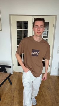 Νέος άνδρας που φοράει καφέ μπλούζα Rip Curl, λευκό παντελόνι και λευκά αθλητικά παπούτσια στέκεται μέσα σε εσωτερικό χώρο μπροστά από πόρτες με γυάλινες επιφάνειες, με το ένα χέρι στην τσέπη.