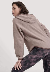 Helle pinke Fleece-Hoodie mit Kapuze, ausgestattet mit einer texturierten Oberfläche und gerippten Bündchen, kombiniert mit dunklen Leggings im lila Batik-Muster.