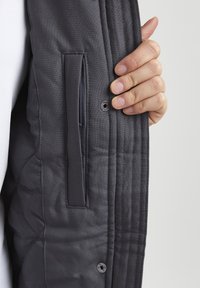 Veste matelassée grise avec un motif de coutures verticales, poche latérale zippée et détail de bouton-pression à l'avant.