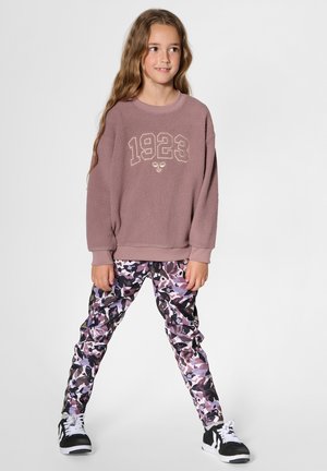 Młoda dziewczyna w mauve polarowej bluzie z napisem „1923” i fioletowych spodniach moro, stoi z rozstawionymi nogami na jednolitym tle.