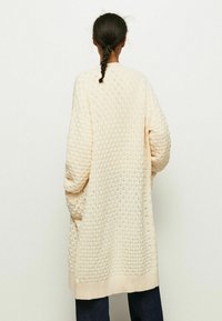 Dlouhý kardigan s otevřeným předním dílem v krémovém úpletu s texturovaným diamantovým vzorem. Má široké rukávy a žebrovaný lem, doplněný bočními kapsami.