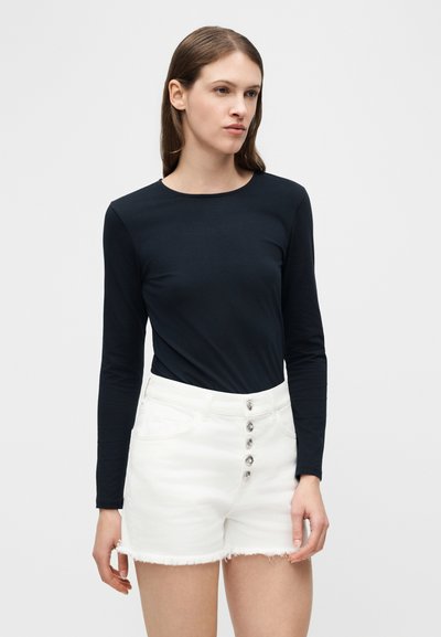 Γυναίκα με ίσια καστανά μαλλιά φοράει εφαρμοστή μαύρη μπλούζα με μακριά μανίκια και ψηλόμεσα λευκά σορτς με κουμπιά, σε απλό φόντο.