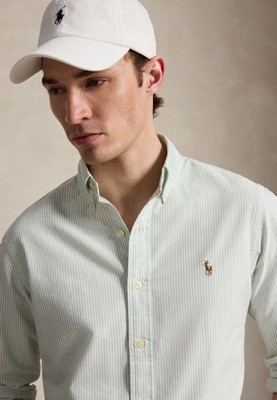 Jeune homme portant une casquette blanche de baseball et une chemise rayée vert clair à boutons avec un logo de joueur de polo sur la poitrine.