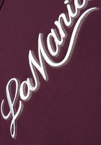 Bordová tkanina s bielym a hnedým tieňovaným vyšitým nápisom „LacMadnie“ v kurzíve diagonálne naprieč povrchom.