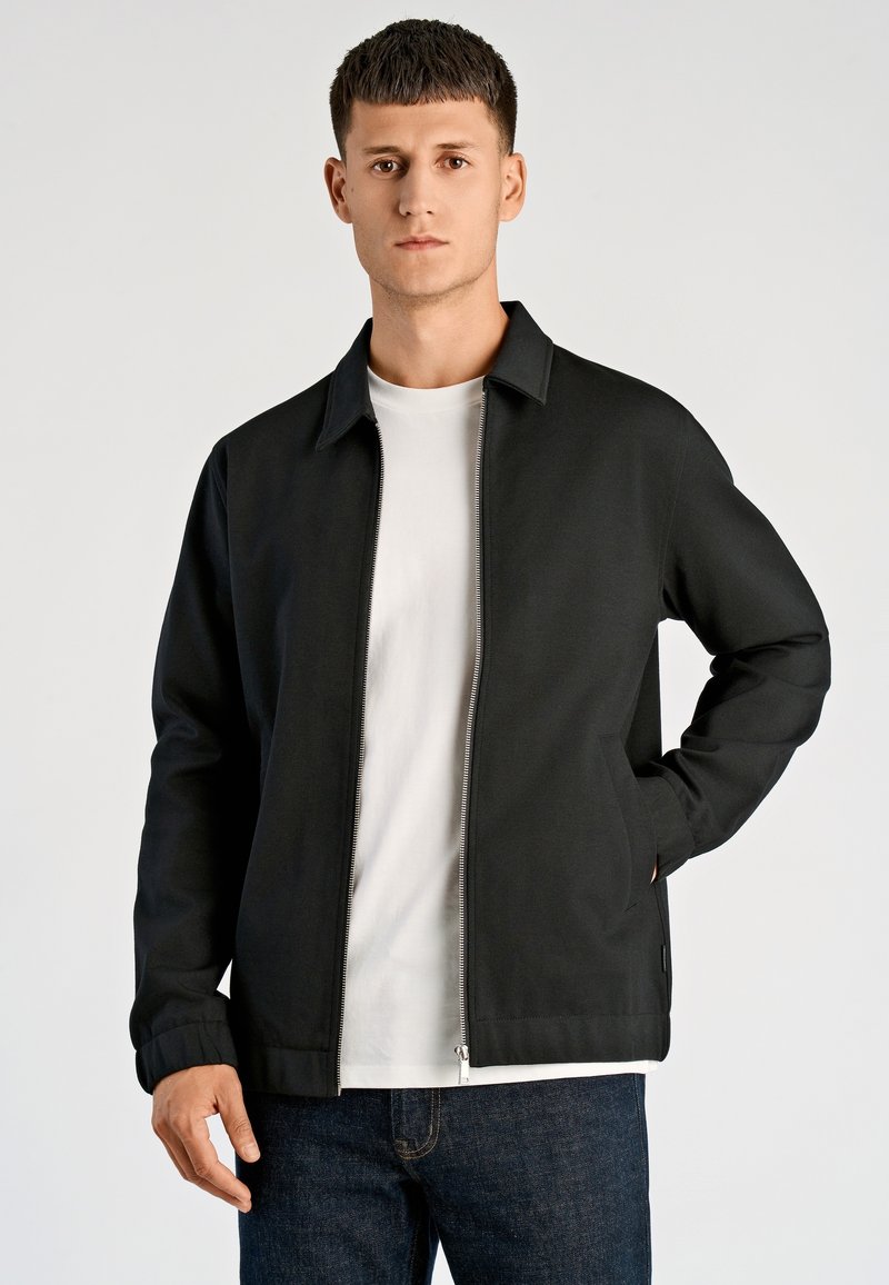 Czarna kurtka zapinana na zamek z ostrym kołnierzem, dwiema bocznymi kieszeniami, ściągaczami na mankietach i gładką fakturą, noszona na białej koszulce.