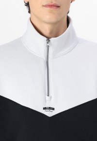 Half-zip tröja i vitt och svart; mjukt tyg med en strukturerad känsla. Har en kontrasterande V-formad design och logotypen på bröstet.
