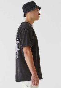 T-shirt en coton noir avec un design graphique violet au dos, assorti à un chapeau seau noir. Vue de profil.