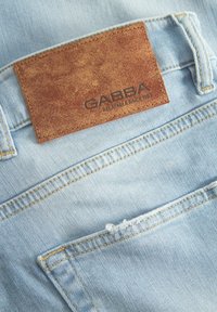 Tejido de mezclilla azul claro con un parche de cuero marrón etiquetado "GABBA". Presenta detalles de costura y un borde deshilachado en el bolsillo.