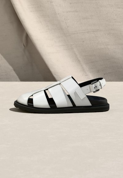 Λευκό δερμάτινο σανδάλι ψαρά με μαύρη σόλα και ρυθμιζόμενο λουράκι με αγκράφα, εμφανίζεται σε πλευρικό προφίλ πάνω σε μπεζ υφασμάτινο φόντο.