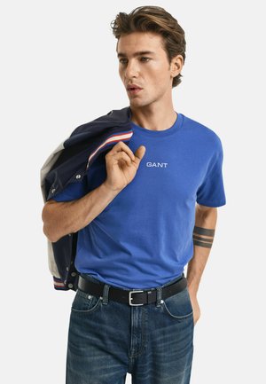 Camiseta de algodón azul real con el logo blanco de "GANT", combinada con jeans de mezclilla oscura y una chaqueta azul marino colgada del hombro. Cinturón negro visible.