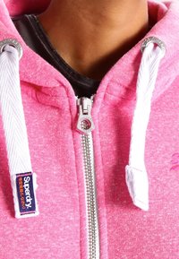Rosa zip-up hoodie i mjukt tyg, med vita dragsko och silverfärgad dragkedja. "Superdry"-logotypen på vänster sida.