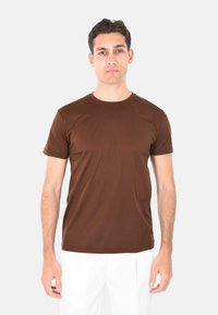 T-shirt marrone a maniche corte realizzato in tessuto morbido, con scollatura rotonda e forma aderente, indossato con pantaloni bianchi.