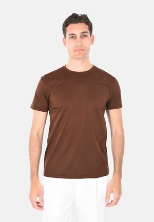 T-shirt marrone a maniche corte realizzato in tessuto morbido, con scollatura rotonda e forma aderente, indossato con pantaloni bianchi.