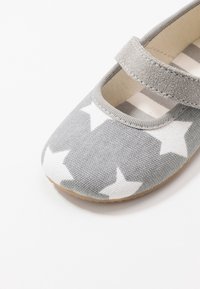Harmaat Mary Jane -kengät, joissa on valkoinen tähtikuosi, kangasremmi ja vaaleanruskea pohja valkoista taustaa vasten.
