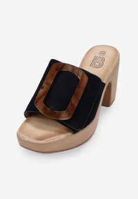 Sandalía de tacón alto de ante negro con una gran hebilla de carey y una suela de plataforma de madera natural, con una textura lisa y un diseño redondeado.