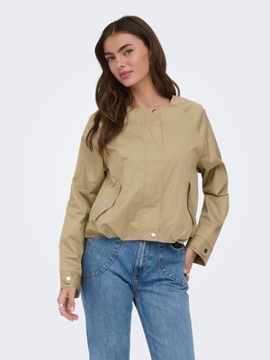 Hosszú barna hajú nő bézs színű, gombos dzsekit és kék farmert viselve, egyik kezét a zsebében tartva, egyszínű háttér előtt áll.