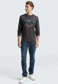 Männliches Model trägt ein dunkelgraues Langarmshirt von Tom Tailor, blaue Jeans und weiße Sneaker und steht vor einem hellgrauen Hintergrund.