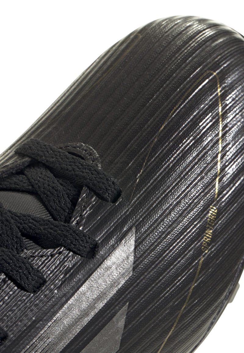 Zapato de fútbol negro con una superficie texturizada y acanalada, que presenta cordones negros y un brillante detalle plateado en el costado. Detalles en dorado presentes.