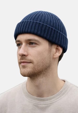 Young man with light beard wearing a navy blue ribbed knit beanie and a light beige crew neck shirt, looking slightly left.