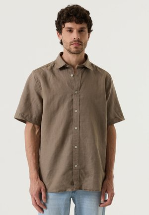 Homme portant une chemise marron à manches courtes boutonnée et un jean bleu clair, debout devant un fond clair uni.