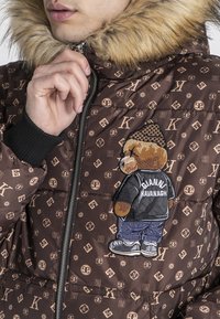 Giubbotto imbottito marrone con cappuccio in pelliccia sintetica, con ricamo di un orso su una giacca nera e jeans blu, accentuato da un motivo ripetuto del logo.