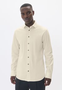 Világos bézs, hosszú ujjú ing, gombos gallérral, barna gombokkal és sima textúrával. Egy modellen látható, semleges háttér előtt.