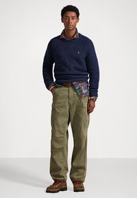 Tengerészkék kötött pulóver kockás ing fölött, olíva színű cargo nadrág és barna csizma piros fűzővel, laza szabású és több zsebjel.