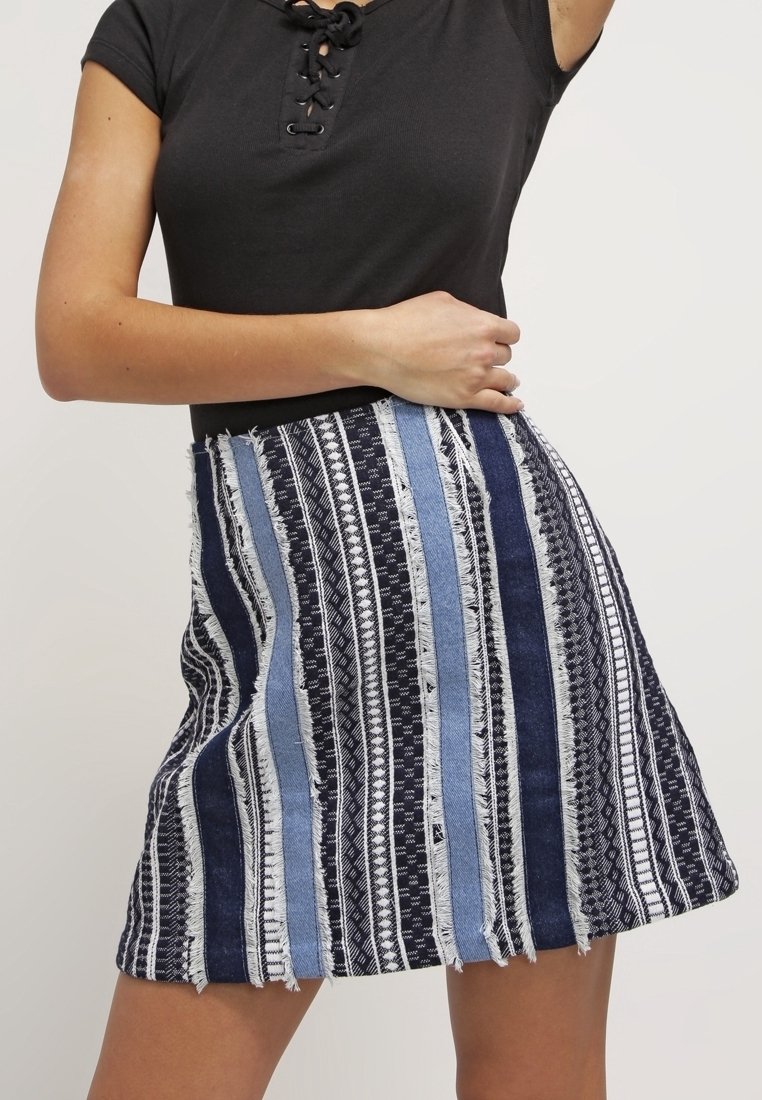 Femme portant un haut noir à lacet au décolleté et une jupe évasée à rayures verticales bleu, noir et blanc texturées, debout avec le bras plié.