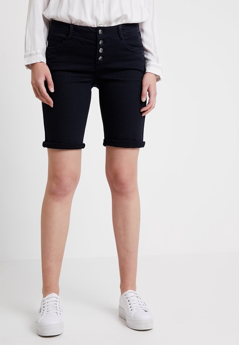 Tmavě modré džíny šortky s přiléhavým střihem, pětikejkový zip, ohrnuté manžety a přední kapsy, v kombinaci s bílými teniskami.