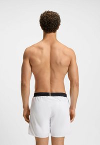 Hátulról látható férfi, göndör hajjal, póló nélkül, fehér fürdőnadrágot visel, fekete derékpánttal, egy sima világos háttér előtt.