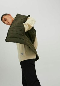 Gestepptes olivgrünes Puffer-Weste mit Reißverschluss vorne, getragen über einem cremefarbenen Strickpullover, in einer dynamischen Pose präsentiert, mit Fokus auf Textur und Schichtung.