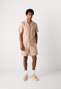 Camisa de manga corta beige y pantalones cortos a juego, con un estampado sutil, cierre de botones, bolsillo en el pecho y combinados con zapatillas color crema.