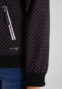 Černá bunda s růžovými puntíky, s kapsou na zip, žebrovanými manžetami a nášivkou Ragwear, ukazující hladkou, lehkou texturu látky.