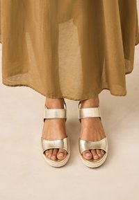 Sandales compensées en cuir doré avec larges brides et fermetures à la cheville. Finition texturée et semelles de style espadrille. Jupe beige clair partiellement visible.