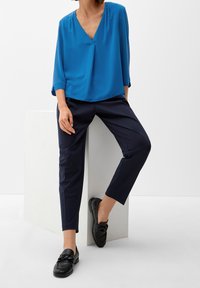 Femme en blouse bleue à col en V et pantalon slim bleu marine appuyée sur un bloc blanc, portant des mocassins noirs, posant devant un fond blanc.