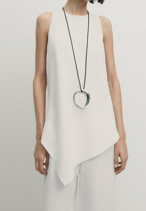 Asymmetrisk vit ärmlös topp i lätt tyg, med en silverfärgad rund hängen på ett svart snören.