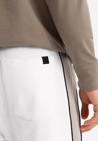Haut à manches longues marron clair associé à un pantalon de survêtement blanc avec une poche arrière et une étiquette noire. Le pantalon a une bande latérale noire.