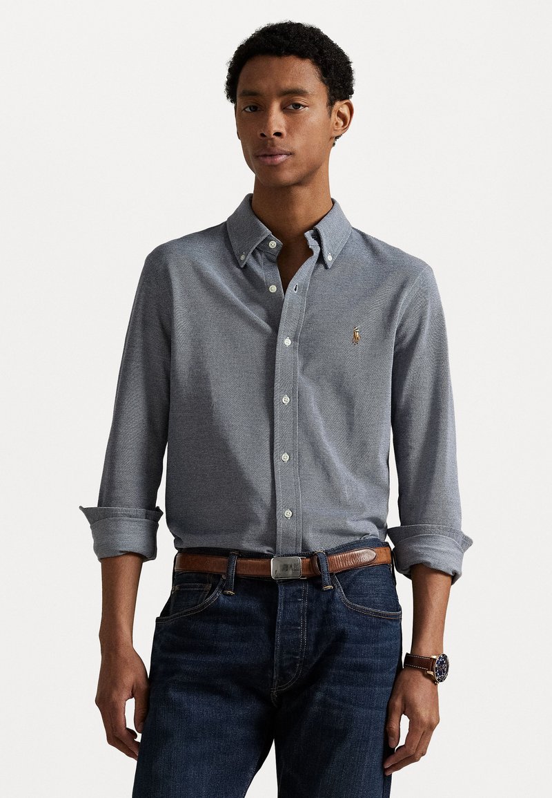 Gray button-down shirt with a textured pattern, long sleeves rolled up, paired with dark jeans and a brown belt. Features a logo on the chest.