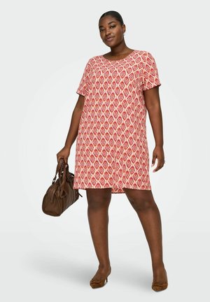 Kvinde iført en kortærmet rød og hvid kjole med geometrisk mønster, holder en brun håndtaske og har brune hæle på, stående selvsikkert.