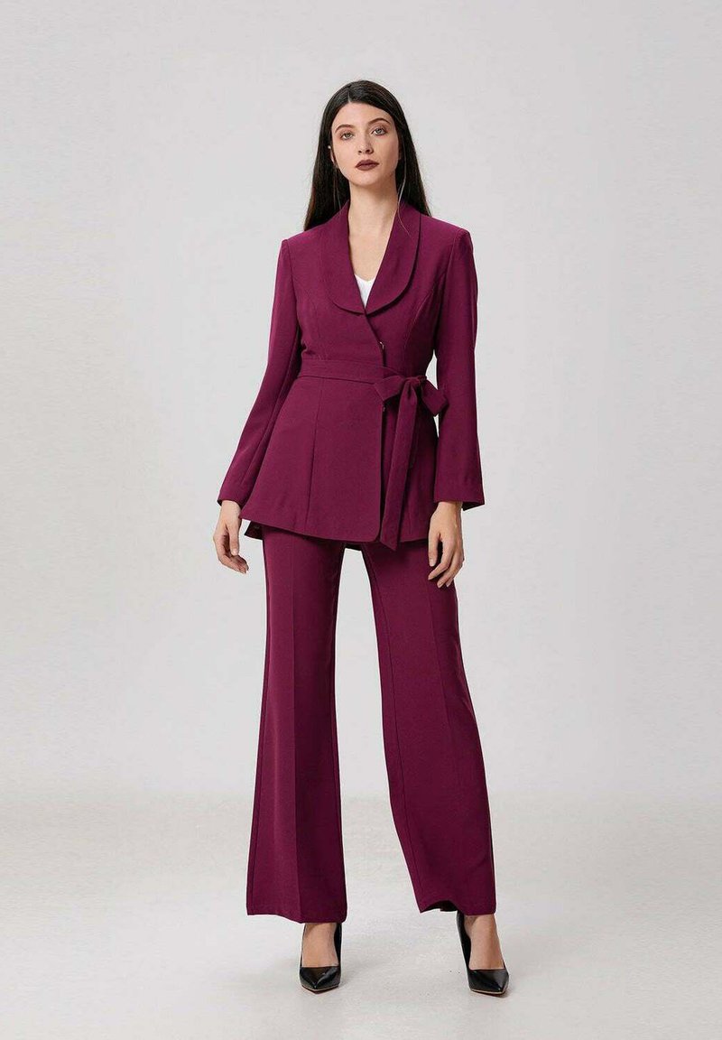 Femme debout portant un tailleur bordeaux avec un blazer à taille liée et un pantalon évasé, associé à des escarpins noirs.