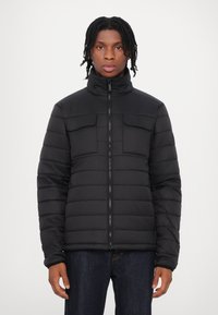 Giacca a vento nera con colletto alto, due tasche sul petto, design trapuntato e zip frontale. Indossata sopra jeans scuri.