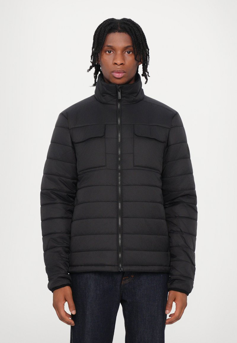 Giacca a vento nera con colletto alto, due tasche sul petto, design trapuntato e zip frontale. Indossata sopra jeans scuri.